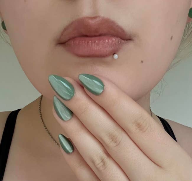 A idiosyncratic with a labret piercing displays a manicure with almond-shaped nails successful a metallic shade, touching their lips