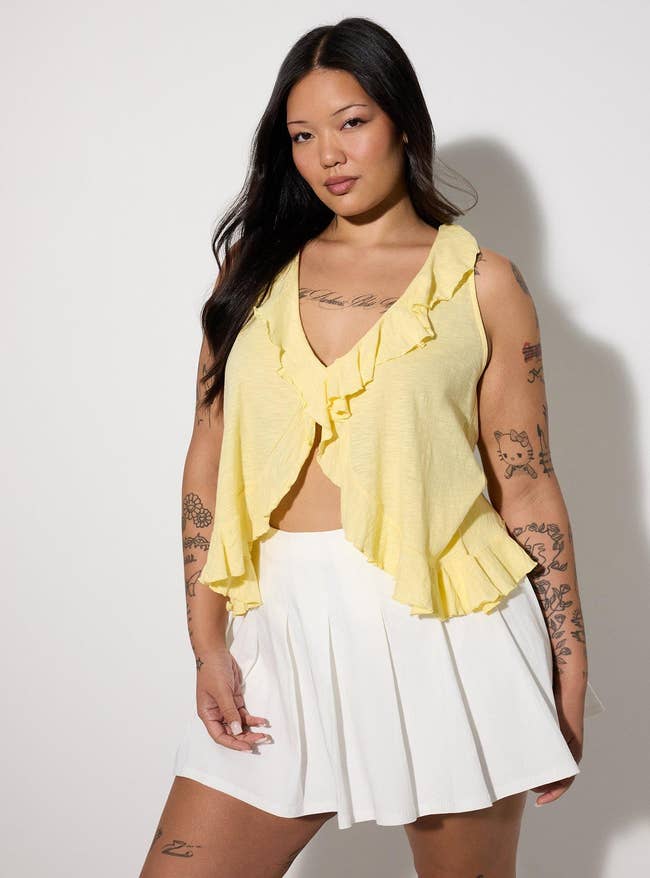 Model wearing a ruffled sleeveless apical  and pleated skirt, featuring tattoos connected  arms and legs, styled for a casual yet chic look
