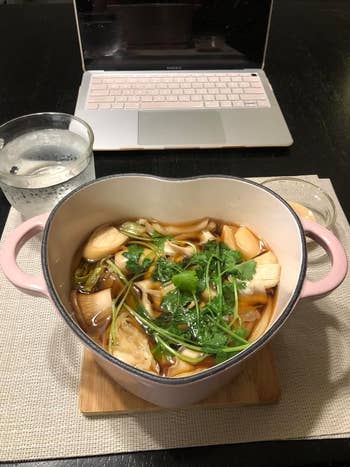 Heart-shaped pot of vegetable soup next to a laptop and glass of water on a table