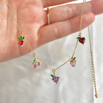 a hand holding a gold necklace with five fruit charms on it