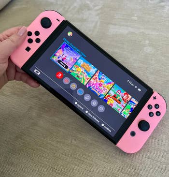 Hand holding a Nintendo Switch with games on screen, pink controllers attached