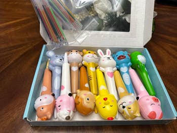 A box of cute animal-themed pens displayed on a wooden table, with a variety of expressions and designs, perfect for adding charm to writing tasks