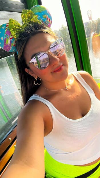 Woman wearing Mickey Mouse ears headband and sunglasses, white top, taking a selfie