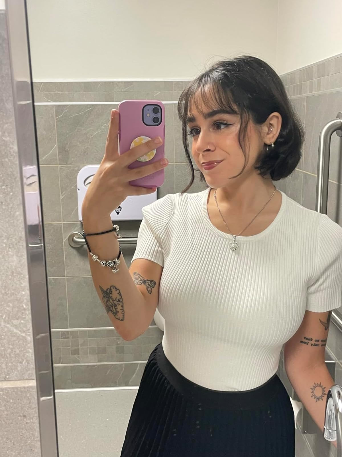 Person taking a mirror selfie, wearing a white ribbed top and black pleated skirt, with tattoos visible on arms