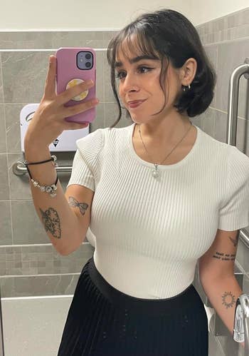Person taking a mirror selfie, wearing a white ribbed top and black pleated skirt, with tattoos visible on arms