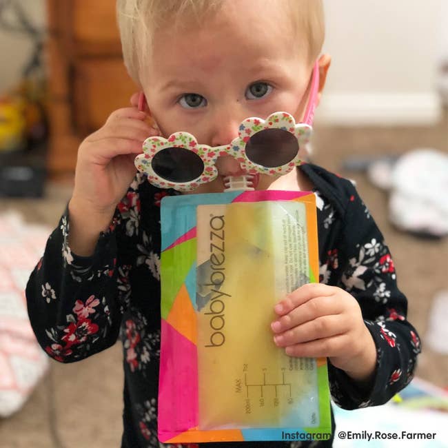 Toddler successful floral sunglasses holds a babe nutrient pouch with a colorful design, lasting successful a country with a playful expression