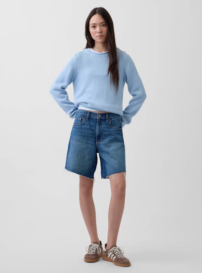 Person wearing a airy  sweater and denim shorts with sneakers, posing with hands connected  hips