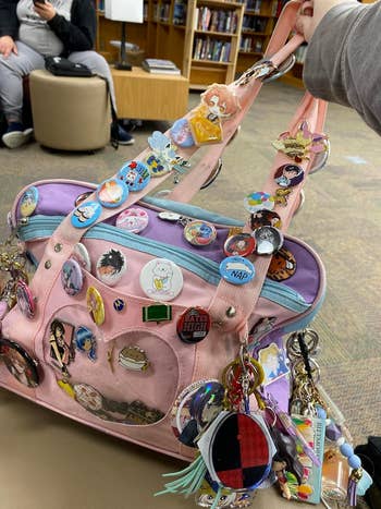 A hand holds a pink and purple bag adorned with anime pins, keychains, and charms