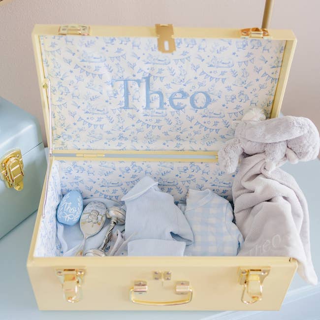 Open decorative container with babe items including clothes, a toy, and tiny gifts, personalized with