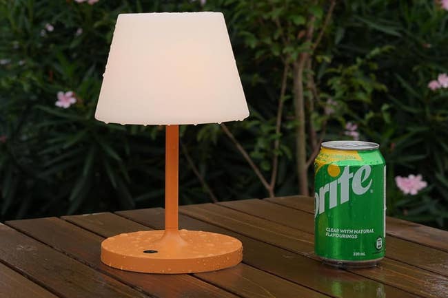 Portable brown lamp wet on an outdoor table next to a can of Sprite showing it's twice the height 