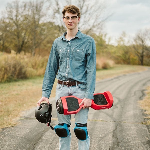 Model on the road holding red hoverboard and helmet while wearing knee pads