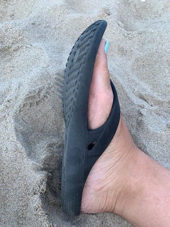 side view of reviewer's foot wearing the black flip-flop in the sand to show its flexible sole