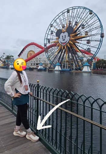 Person posing by a fence with a Mickey Mouse-themed Ferris wheel in the background. Arrow points to their sneakers