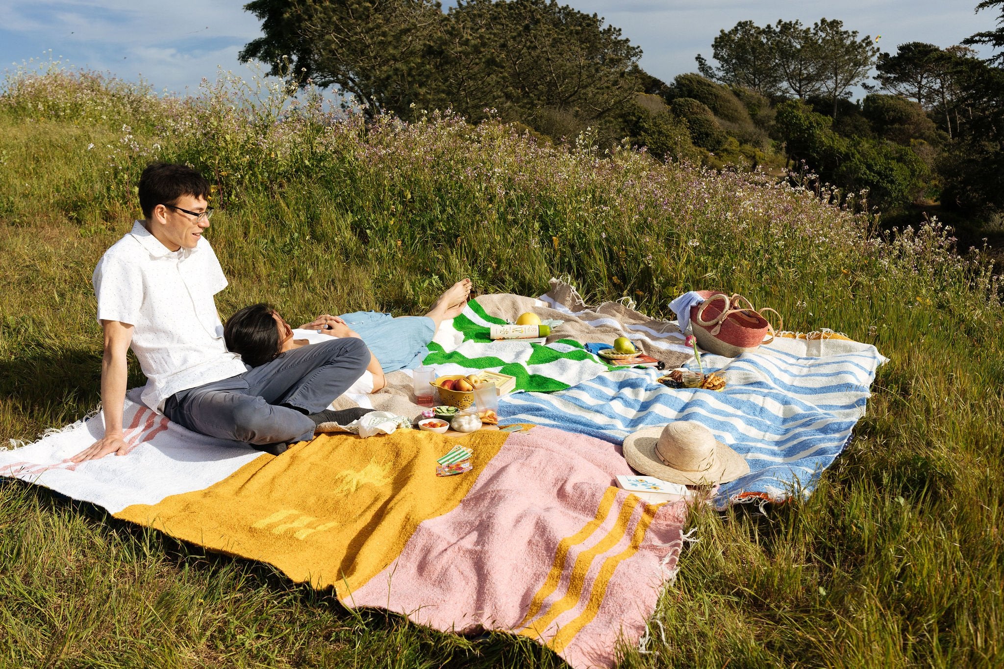 two blankets placed on top of each other outside. a couple sits on top of them with snacks and accessories scattered around.