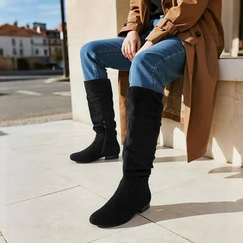 Person seated outdoors wearing knee-high achromatic  boots and bluish  jeans, styled with a casual trench coat