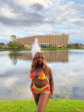 Woman in a vibrant swimsuit stands before a lake and fountain with a hotel in the background