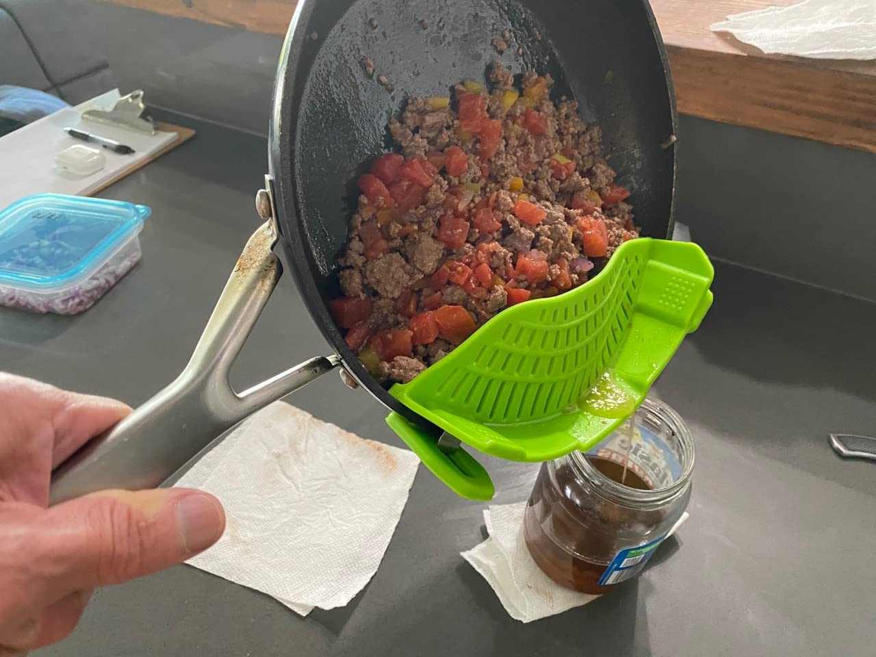 Ground beef and tomatoes are being drained from a skillet into a jar using a bright green strainer clip