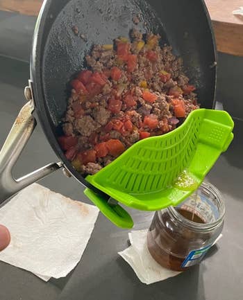 Ground beef and tomatoes are being drained from a skillet into a jar using a bright green strainer clip