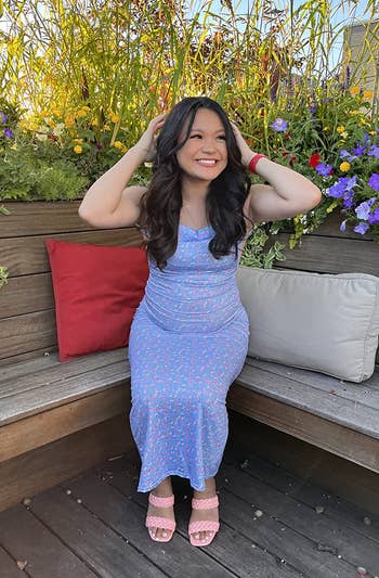 Woman successful a floral formal sitting connected a woody seat surrounded by flowers, smiling and holding her hair