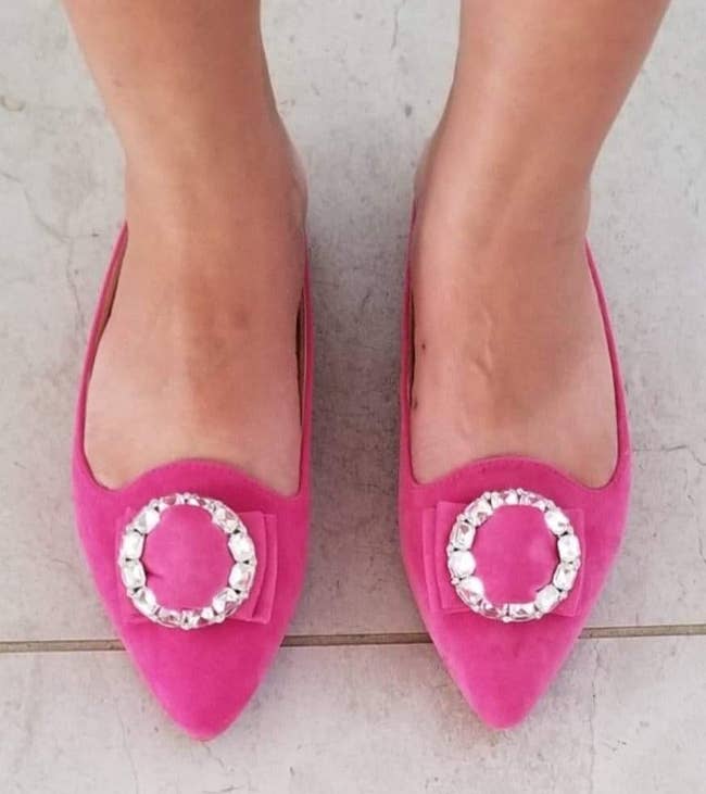 reviewer wearing the pointed-toe slip-on flat with pointed toe in pink suede-like fabric and circular embellishment on top 