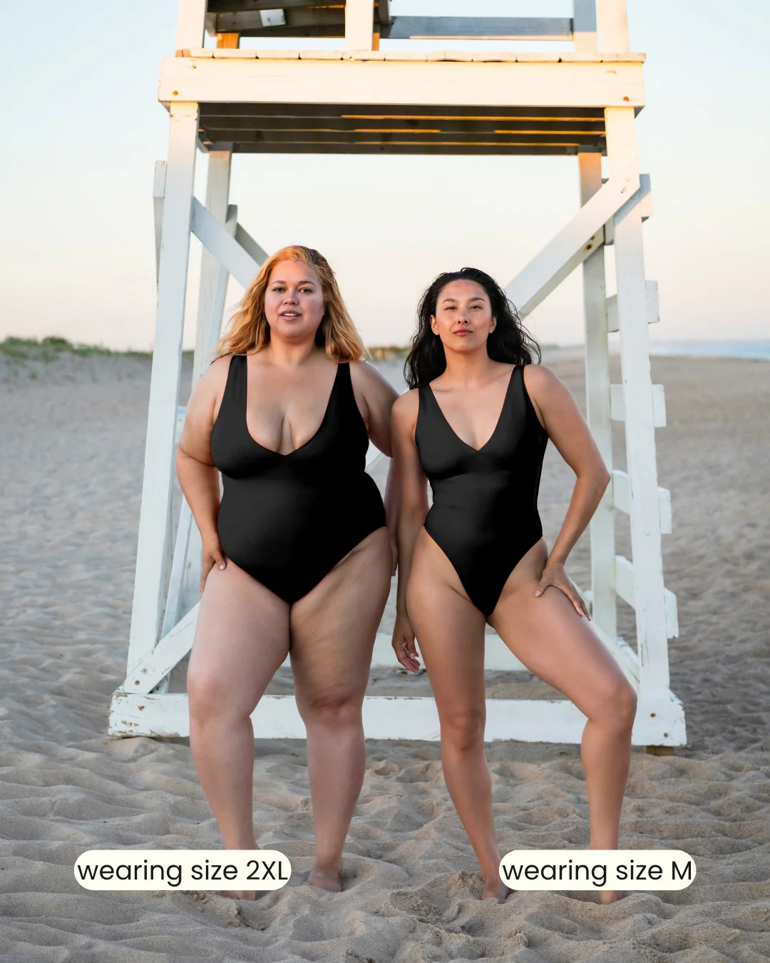 Two women on a beach wear black swimsuits in sizes 2XL and M, standing confidently by a lifeguard stand