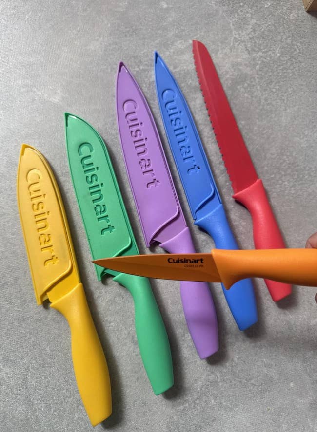 Set of six Cuisinart knives with colorful sheaths, displayed connected a countertop, each matching its respective weapon handle