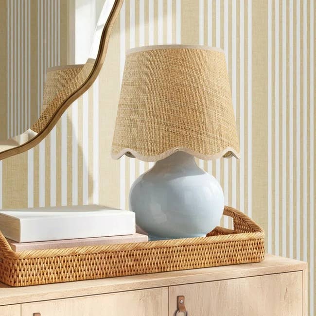 A ceramic table lamp with a woven rattan shade sits on a wooden dresser with a rectangular mirror and stacked books nearby