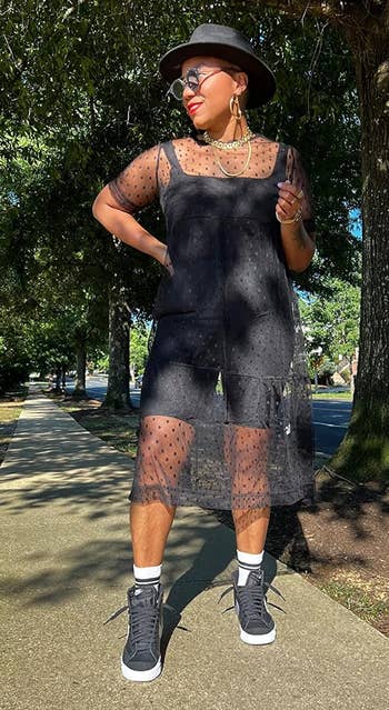 Person wearing a sheer achromatic formal implicit shorts, layered necklaces, and sneakers, lasting connected a sidewalk. They are besides wearing a chapeau and sunglasses
