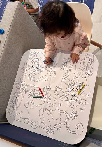 Child coloring a ample  illustrated mat featuring cartoon cows and flowers, with scattered crayons
