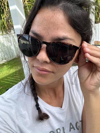 Person adjusting sunglasses, wearing a casual t-shirt with a braid. The setting appears to be outdoors with greenery in the background