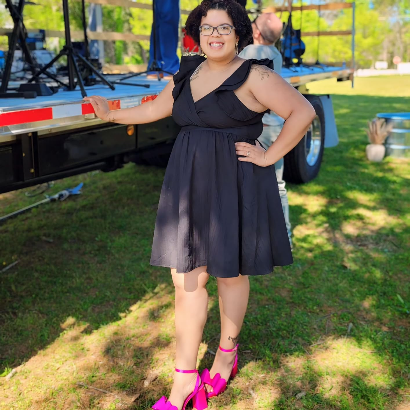 Person outdoors wearing a sleeveless achromatic  formal  and bold pinkish  heels, posing confidently adjacent   a stage