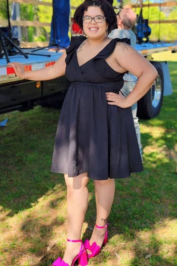 Person outdoors wearing a sleeveless achromatic formal and bold pinkish heels, posing confidently adjacent a stage