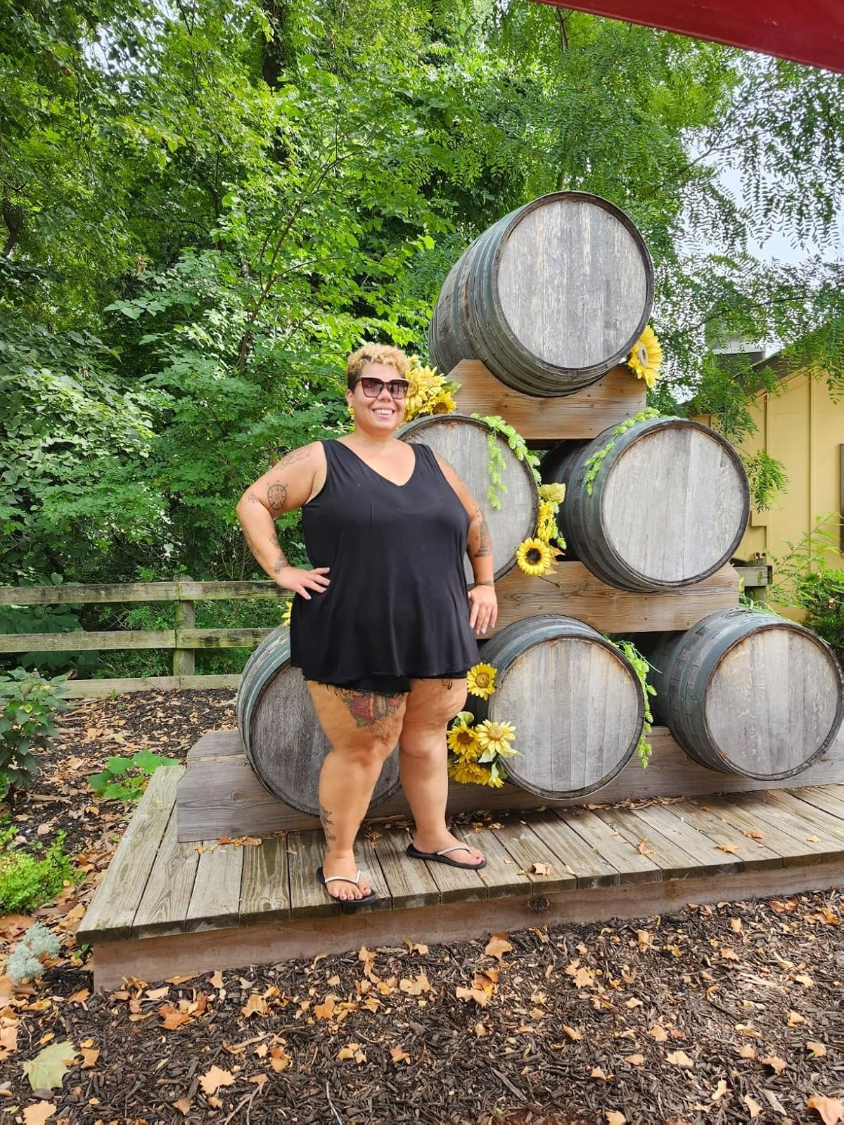 Plus-size idiosyncratic   successful  a sleeveless achromatic  formal  and sandals stands smiling successful  beforehand   of a stack of ample  woody  barrels adorned with sunflowers