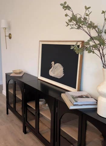 a framed swan people connected a achromatic console table, with a vase of branches and stacked books, creating an elegant setting