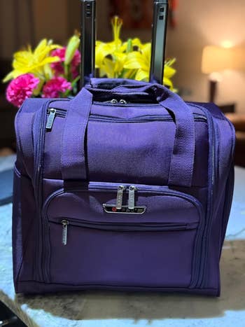 Travel-sized, purple rolling luggage with front pockets, displayed on a marble surface. Yellow and pink flowers are visible in the background