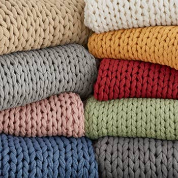 Neatly stacked, chunky-knit blankets in various colors. Perfect for cozying up and adding texture to home decor. Ideal for shopping enthusiasts looking for stylish blankets