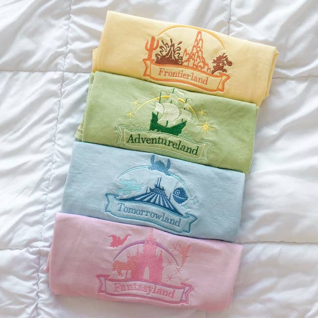 four different colored tees with embroidered disney lands on them from within magic kingdom