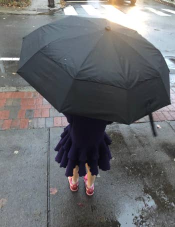 Person with an umbrella wearing a ruffled skirt and reddish  shoes stands connected  a bedewed  sidewalk adjacent   a crosswalk