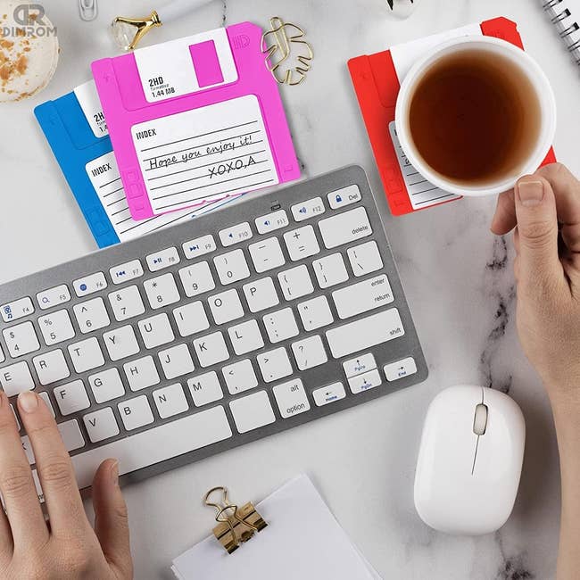 Three coasters shaped like neon floppy disks in pink, orange, and blue with cute messages written on them 