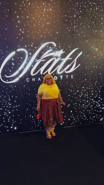 Person successful casual attire with a agleam yellowish apical and plaid skirt stands smiling against a starry backdrop astatine Stats Charlotte