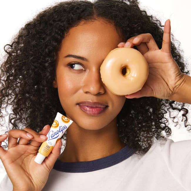 Person playfully holds a glazed donut implicit 1 oculus and a Lanolips articulator balm, highlighting a amusive attack to quality products