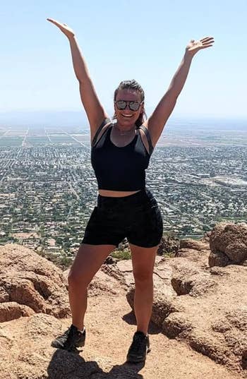 A person poses with arms up on a mountaintop overlooking a vast cityscape, wearing sporty attire suitable for hiking