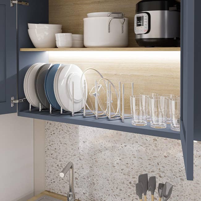 Kitchen cabinet with organized plates, bowls, and glasses on shelves; an electric cooker on the top shelf. Spatulas in a holder near the sink