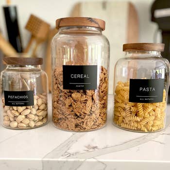Three labeled pantry jars containing pistachios, cereal, and pasta, arranged by size on a countertop