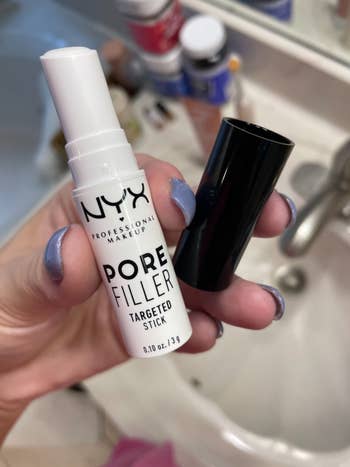 Hand holding NYX Pore Filler Targeted Stick adjacent to a achromatic cap, with a descend disposable successful the background