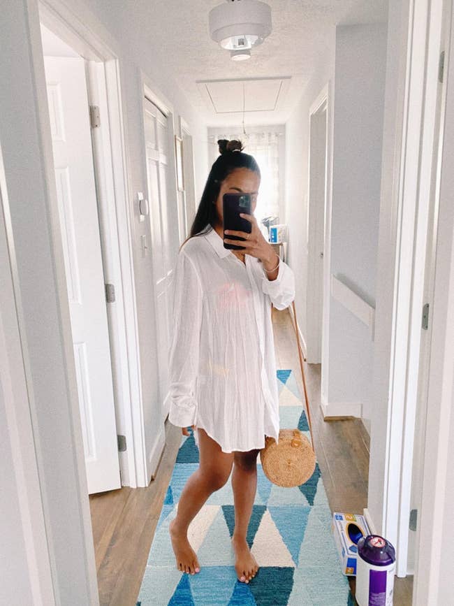 Person taking a reflector  selfie successful  a hallway, wearing an oversized garment  and holding a circular  straw bag. Barefoot connected  a geometric rug