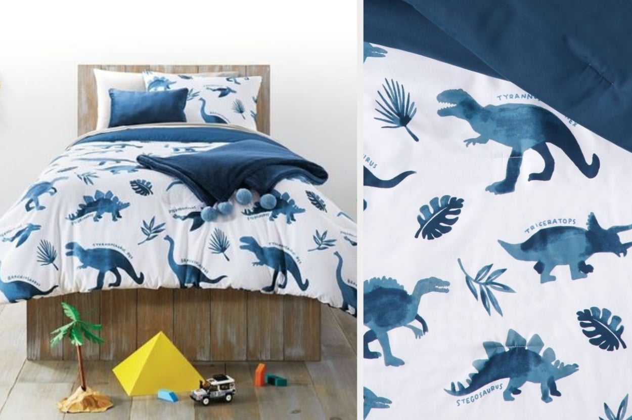 Blue and white dinosaur watercolor patterned comforter with matching pillow next to navy blue pillow and solid blue throw blanket with pom poms on a wooden bedframe, close up of comforter design with navy blue underside