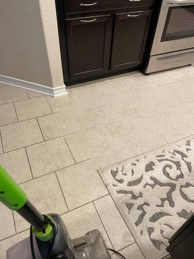 Vacuum cleaner connected a tiled room level adjacent cabinets and an oven. A patterned rug is partially visible