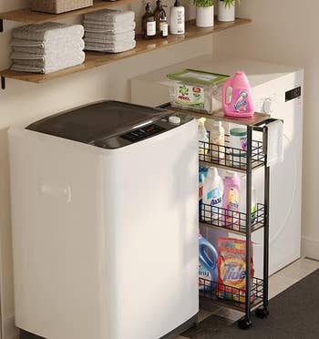 Top-loading washing machine next to laundry supplies on a shelf and a rolling cart filled with detergent and fabric softener