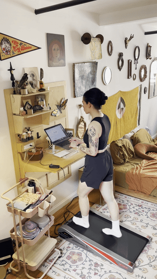 Person in sportswear using a laptop on a standing desk with a treadmill in a room with eclectic decor